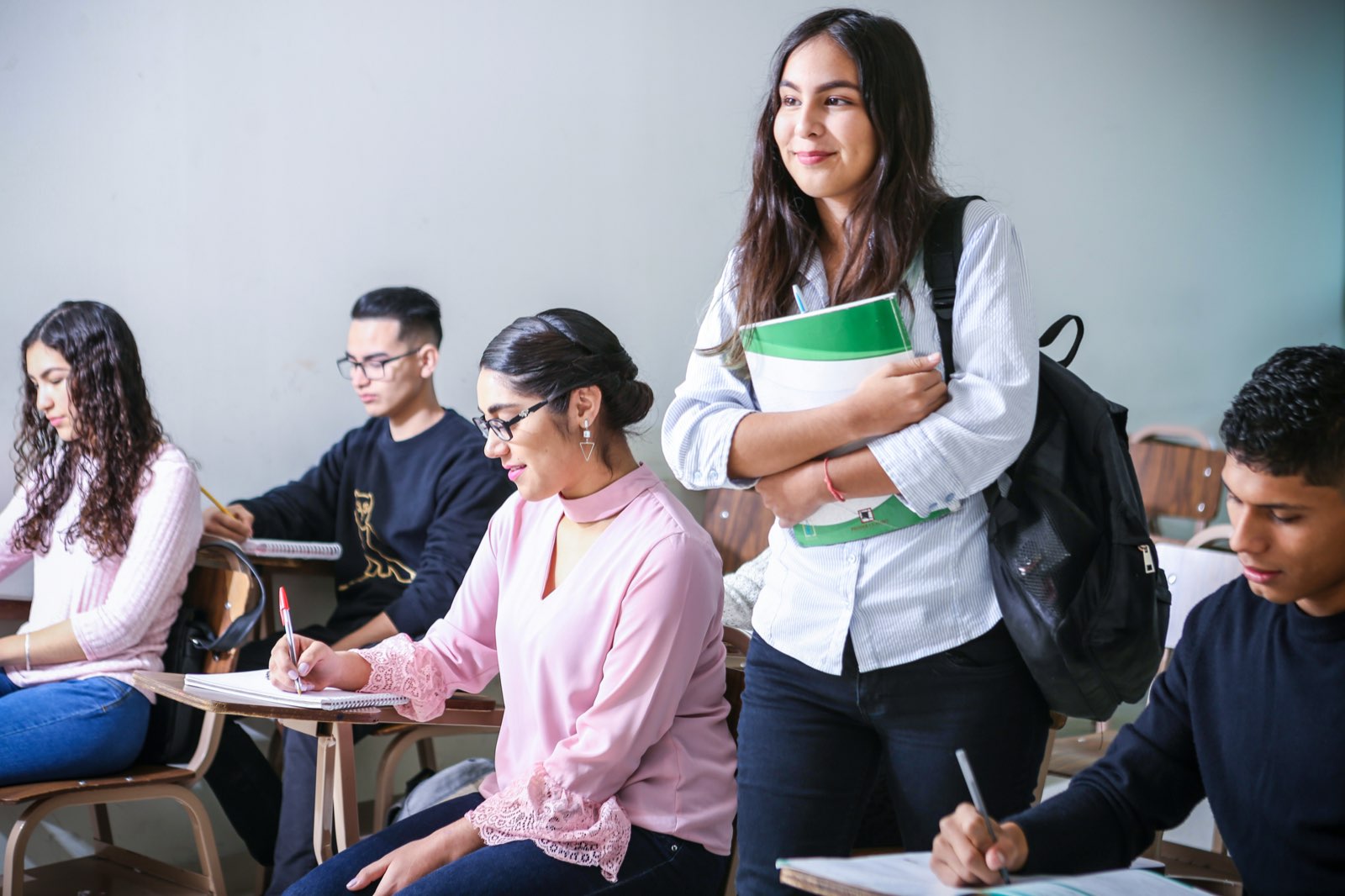 Students in a classroom; one stands holding her notebooks, smiling.