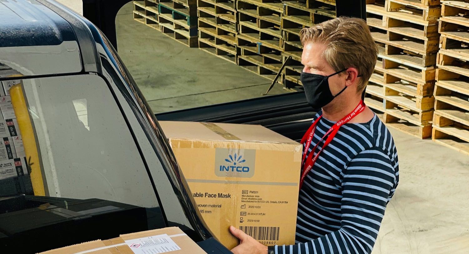 A warehouse worker loads a carton into a vehicle at a dispatch bay, with stacked pallets behind him.